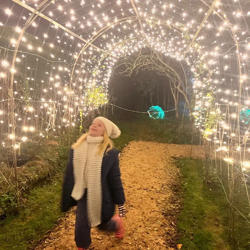 A child in a woolly hat and scarf walking along a gravel path through a tunnel of white fairy lights in the garden at Gilbert White's House