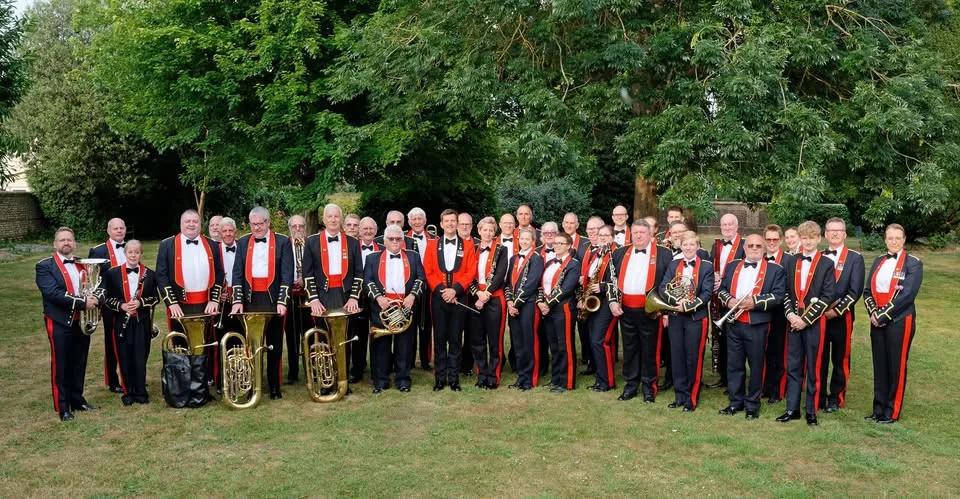 About forty members of the Royal Marines Association Concert Band in black and red uniforms standing on grass in front of trees, several holding tubas, saxophones, a French horn and other brass and woodwind instruments
