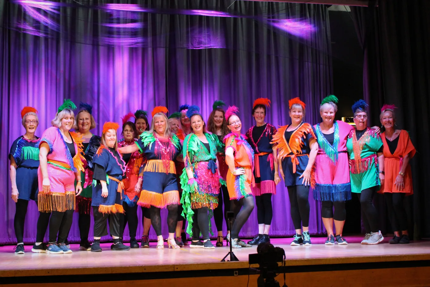 A line of performers on a stage in front of a purple-lit curtain, wearing bright fringed costumes in pink, orange, green and navy with feathered headpieces