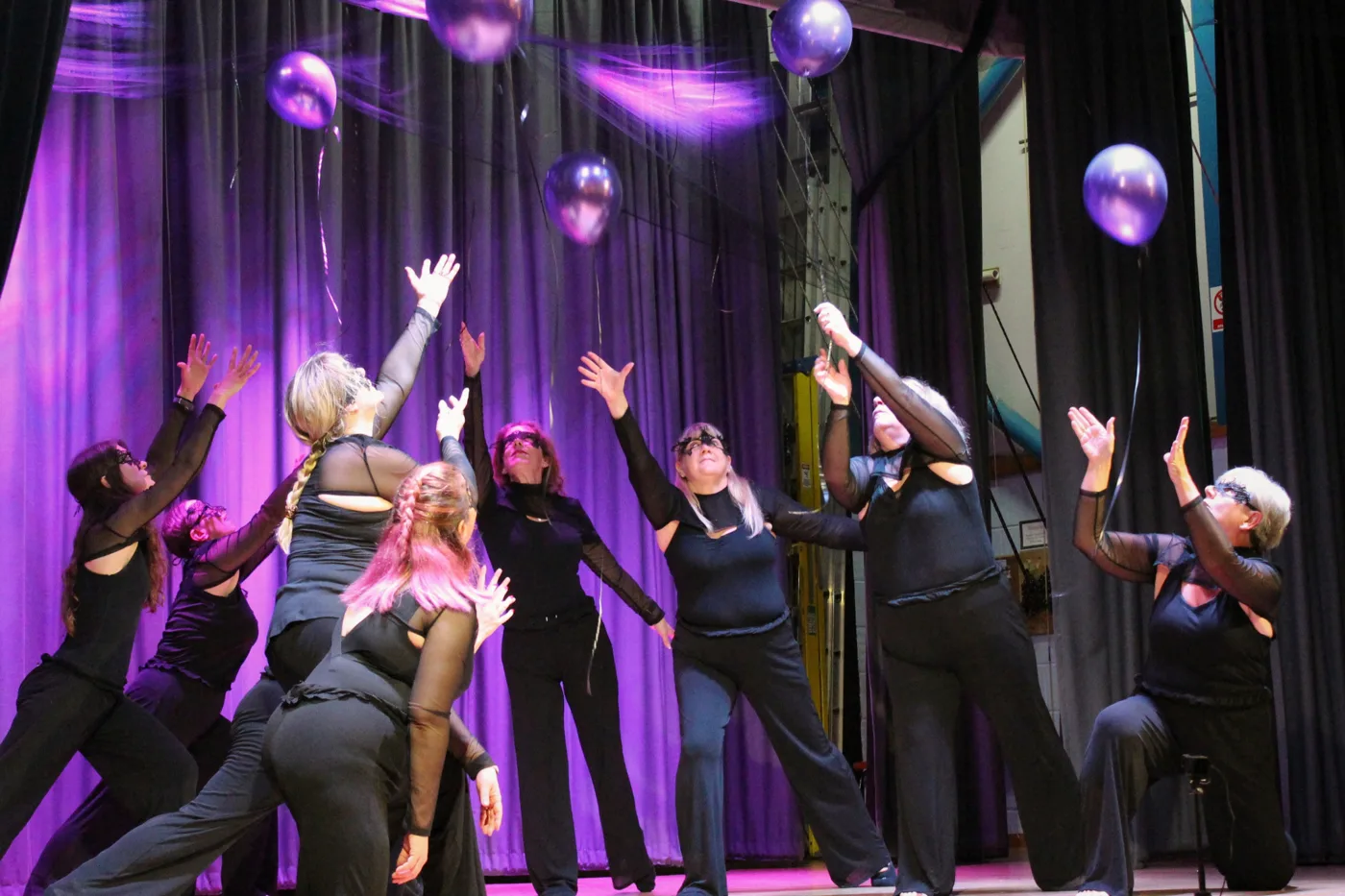 Dancers in black reaching up towards purple balloons floating above a stage lit in purple