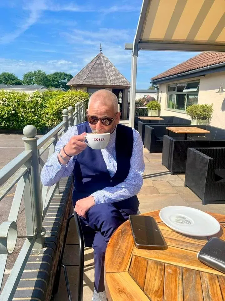 Rob Fielder in sunglasses drinking from a Costa cup at a table on a sunny terrace