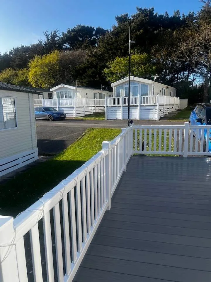 Grey decking with a white rail looking out across grass to two holiday homes and pine trees
