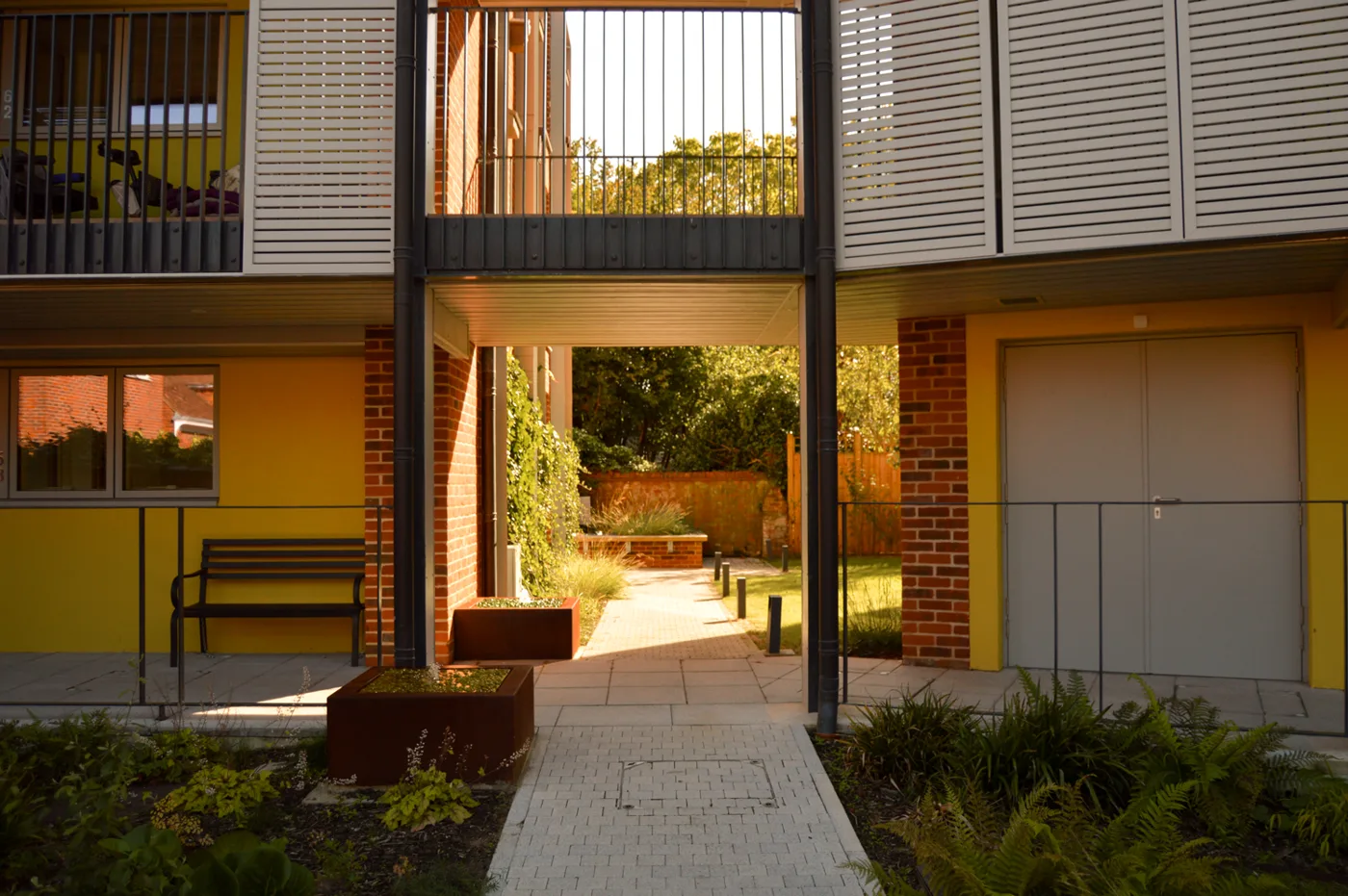 Yellow-walled almshouse flats with balconies and a paved path through to a garden