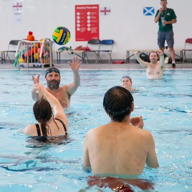 A water polo ball in the air above a group of players in a swimming pool, with a coach standing on the poolside